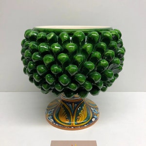 Puede incluir: Un macetero de cerámica verde con forma de piña con una base pintada a mano de colores. El macetero está hecho en Sicilia, Italia.