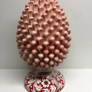 Puede incluir: Una gran escultura de piña de cerámica rosa con una base roja y blanca. La base está decorada con un patrón floral.