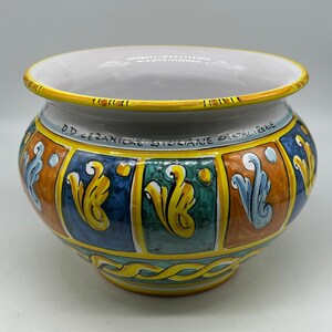 May include: A ceramic planter with a wide, rounded shape. The planter features a white interior and a vibrant exterior design with yellow, blue, orange, and green panels. The text "D.D. CERAMICHE SICILIANE CALTAIRENE" is written around the rim.