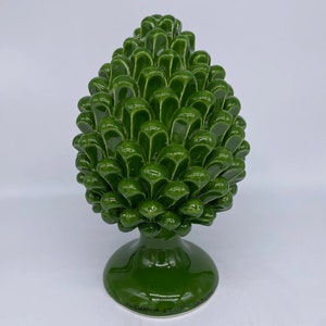 Puede incluir: Un adorno de piña de cerámica verde con base. El adorno está hecho en Italia y tiene el texto "Ceramiche Siciliane" en la base.