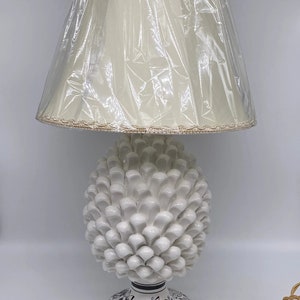 Puede incluir: Lámpara de mesa de cerámica blanca con un diseño de piña. La lámpara tiene una pantalla de tela blanca con un ribete dorado. La base de la lámpara está decorada con diseños florales en blanco y negro. La lámpara está sobre una superficie blanca.