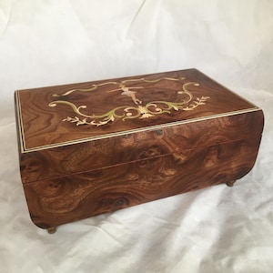 Op de afbeelding: Een rechthoekige houten muziekdoos met een gedetailleerd ingelegd ontwerp op het deksel. De doos heeft een ballerinafiguur en bloemaccenten in goud, groen en wit. Het hout heeft een rijke, wervelende nerf.