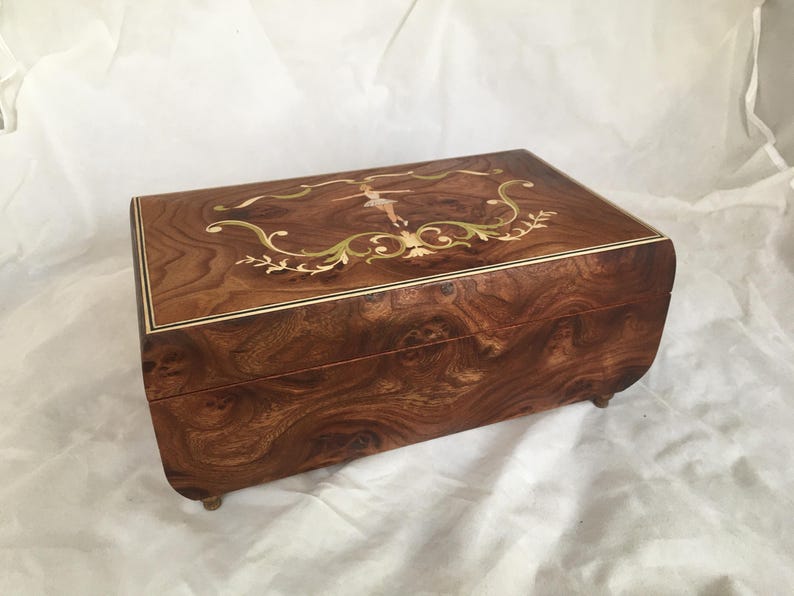 Op de afbeelding: Een rechthoekige houten muziekdoos met een gedetailleerd ingelegd ontwerp op het deksel. De doos heeft een ballerinafiguur en bloemaccenten in goud, groen en wit. Het hout heeft een rijke, wervelende nerf.