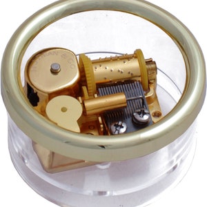 May include: A small, clear, round music box with a gold-colored metal rim and internal components. The mechanism is visible, featuring a rotating cylinder with pins and a comb-like structure. The base is clear plastic, and the music box has the text "N.J. Dean & Co."