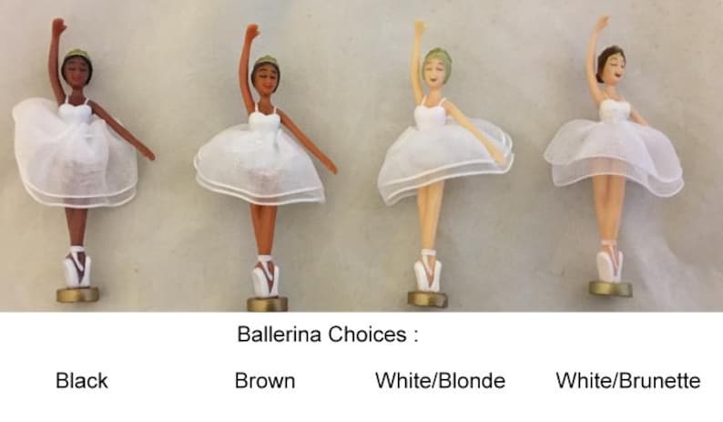 Op de afbeelding: Vier ballerina beeldjes met verschillende huidtinten en haarkleuren. Elk beeldje draagt een witte tutu en spitsschoenen. De tekst "Ballerina Choices" staat eronder, met de labels: Black, Brown, White/Blonde en White/Brunette.