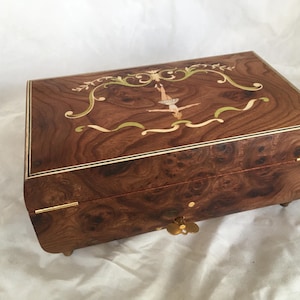 Op de afbeelding: Een rechthoekige houten muziekdoos met een ingelegd ontwerp. De doos heeft een ballerina in het midden, omringd door een sierlijke rand. Het hout is donkerbruin met een uniek nerfpatroon. Goudkleurige hardware en kleine voetjes maken het ontwerp compleet. Ongeveer 20 cm lang.
