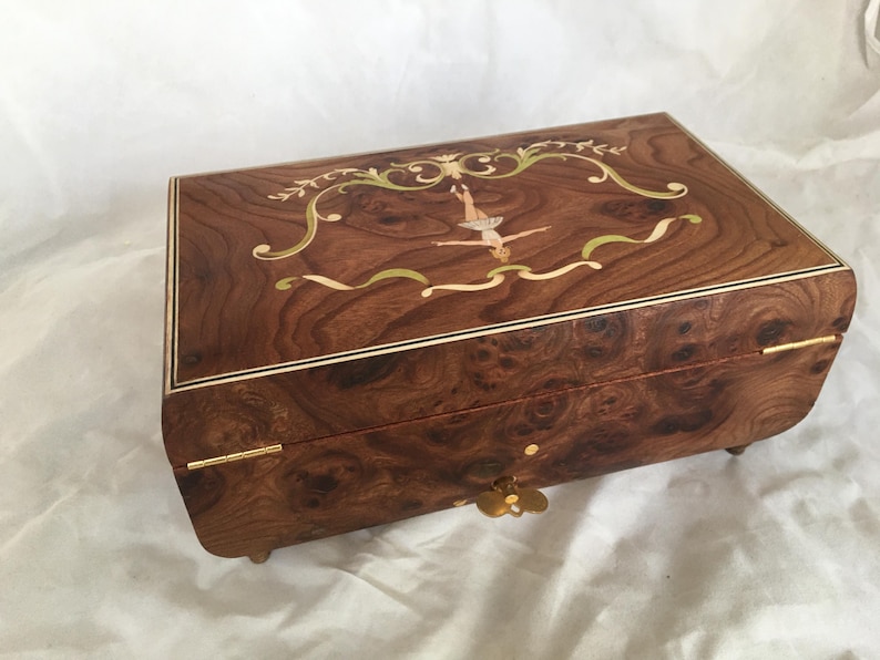 Op de afbeelding: Een rechthoekige houten muziekdoos met een ingelegd ontwerp. De doos heeft een ballerina in het midden, omringd door een sierlijke rand. Het hout is donkerbruin met een uniek nerfpatroon. Goudkleurige hardware en kleine voetjes maken het ontwerp compleet. Ongeveer 20 cm lang.