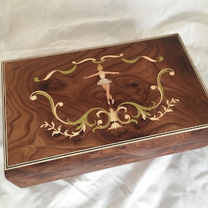 Op de afbeelding: Een rechthoekige houten muziekdoos met een ballerina-inleg. De doos is gemaakt van donkerbruin hout met een decoratieve rand. Het deksel heeft een ballerina in een tutu, omringd door groene en cr&egrave;mekleurige bloemaccenten.
