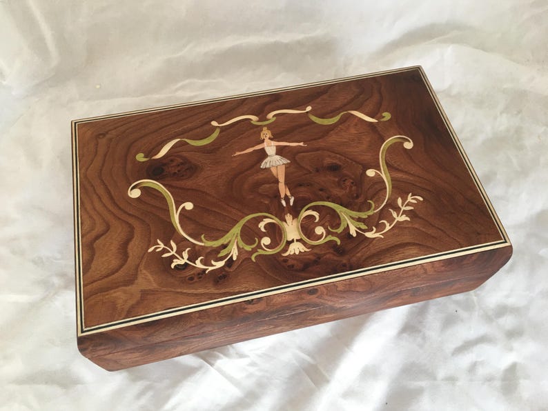Op de afbeelding: Een rechthoekige houten muziekdoos met een ballerina-inleg. De doos is gemaakt van donkerbruin hout met een decoratieve rand. Het deksel heeft een ballerina in een tutu, omringd door groene en cr&egrave;mekleurige bloemaccenten.