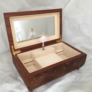Op de afbeelding: Een houten muziekdoos met een spiegelinterieur en een ballerinafiguur. De doos is open en onthult een beige interieur en een kleine ballerina in een witte tutu. De buitenkant is donkerbruin met een houtnerfpatroon.