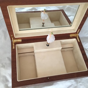 Op de afbeelding: Houten muziekdoos met een spiegeldeksel, met een ballerinafiguur. De binnenkant is bekleed met beige stof. De doos is open en onthult de ballerina en haar weerspiegeling. De scharnieren en sluitingen zijn goudkleurig.