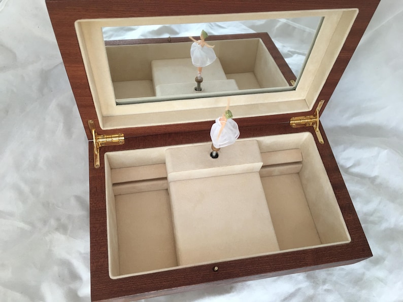 Op de afbeelding: Houten muziekdoos met een spiegeldeksel, met een ballerinafiguur. De binnenkant is bekleed met beige stof. De doos is open en onthult de ballerina en haar weerspiegeling. De scharnieren en sluitingen zijn goudkleurig.