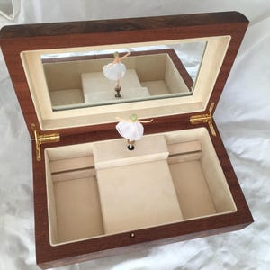 Op de afbeelding: Een houten sieradendoos met een spiegeldeksel en een ballerinafiguur. De doos is open en onthult een beige interieur met compartimenten. De ballerina draagt een witte tutu en staat op een centraal platform. De doos is gemaakt van donker hout.