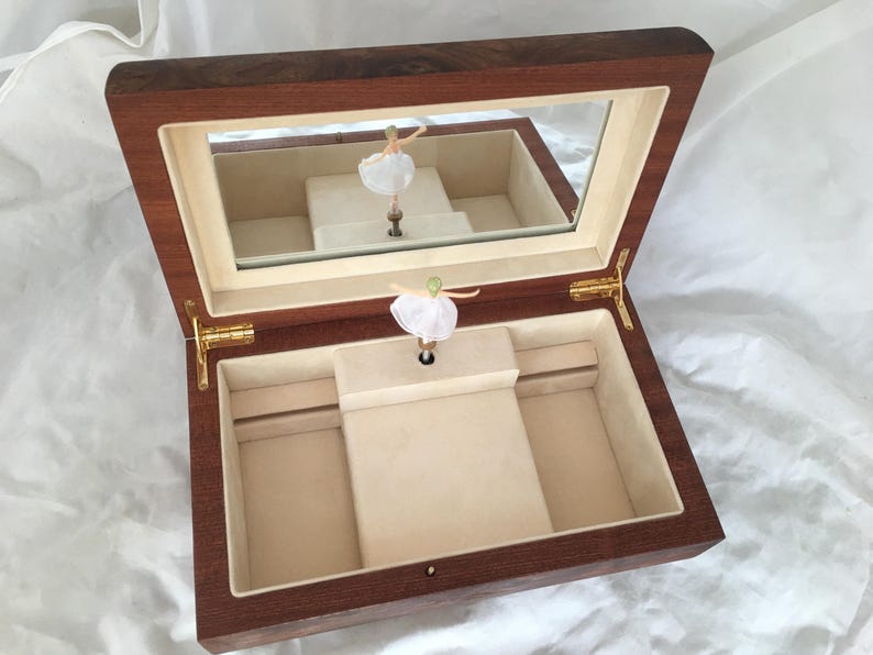 Op de afbeelding: Een houten sieradendoos met een spiegeldeksel en een ballerinafiguur. De doos is open en onthult een beige interieur met compartimenten. De ballerina draagt een witte tutu en staat op een centraal platform. De doos is gemaakt van donker hout.