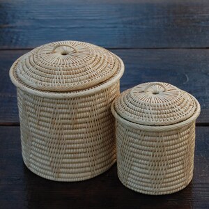 May include: Two woven wicker storage baskets with lids. The larger basket is taller than the smaller one. Both baskets are cylindrical with a natural beige color and a woven pattern. The lids have a circular handle.