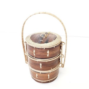 May include: A three-tiered, wooden food carrier with a woven handle. The carrier is made of dark brown wood with woven accents and a light-colored woven handle. The carrier is designed to stack and has a lid.