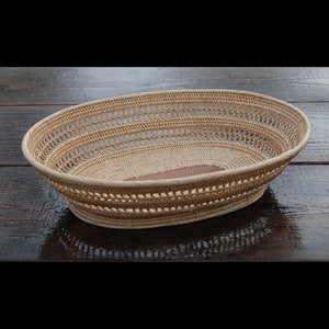 May include: Oval-shaped woven basket with a light brown hue. The basket features an open weave design around the sides and a solid brown base. The basket is made of natural materials and is suitable for storage or display.