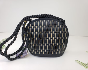 May include: A black and tan woven handbag with a braided black rope strap. The bag has a rounded shape and a woven pattern. The strap is thick and appears to be made of a soft material. The bag is on a white surface.