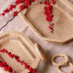 May include: Two woven wicker trays with handles and a diamond pattern are displayed. A sprig of red berries drapes across the trays. Two woven napkin rings are also visible, suggesting a dining or home decor theme.