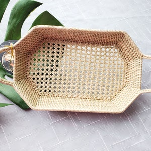 May include: A light beige woven rattan basket with a rectangular base and rounded corners. The basket features an open diamond-patterned weave on the sides and bottom, with two integrated curved handles on opposite ends for easy carrying.