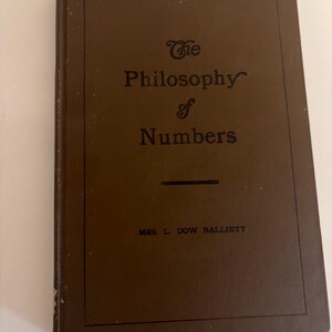 May include: A brown hardcover book titled The Philosophy of Numbers by Mrs. L. Dow Balliett. The front cover features a simple black rectangular border, black text for the title, and the author's name printed in black at the bottom center.