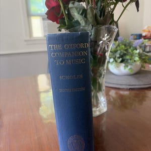 May include: A dark blue hardcover book titled The Oxford Companion to Music by Scholes, Tenth Edition. The spine features gold lettering and an Oxford University Press emblem at the bottom. The book stands upright with a clear glass vase of red roses.
