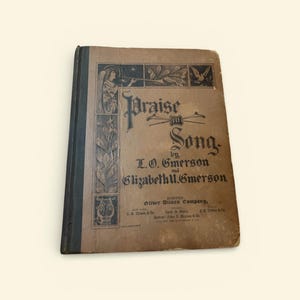 May include: An antique book titled "Praise in Song" by L.O. and Elizabeth U. Emerson. The cover is a faded brown with black decorative elements, including an angel playing a horn. The book is from the Silver, Ditson Company.