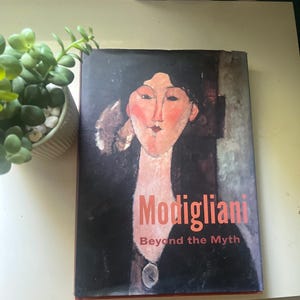 May include: A book titled "Modigliani Beyond the Myth" features a portrait of a woman with a long neck and red cheeks. The book cover is dark with red lettering and a portrait of a woman.