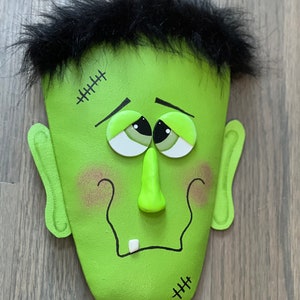 May include: A green Frankenstein face with black hair, stitched scars, and googly eyes. The face is made of felt and has a 3D nose and a single tooth.