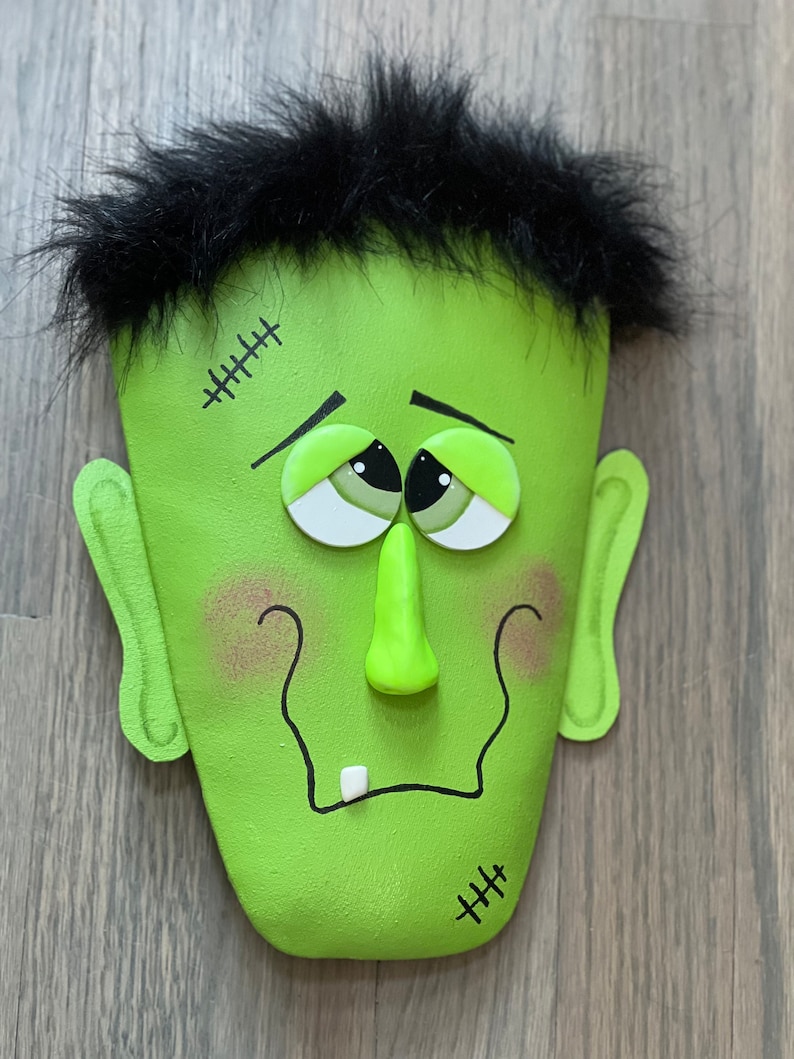 May include: A green Frankenstein face with black hair, stitched scars, and googly eyes. The face is made of felt and has a 3D nose and a single tooth.