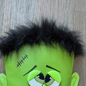 May include: A green monster face with black hair, stitched scar, and large green eyes. The monster has a green nose and a slight smile.
