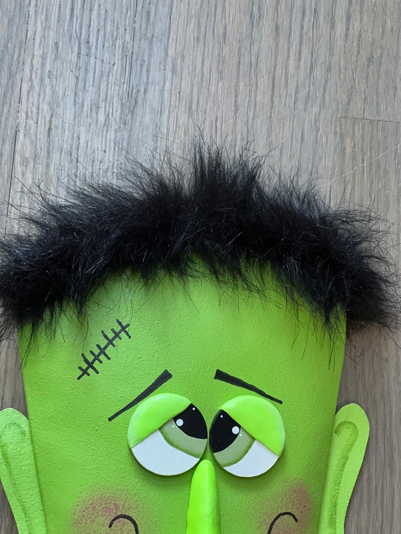 May include: A green monster face with black hair, stitched scar, and large green eyes. The monster has a green nose and a slight smile.