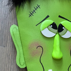 May include: Close-up of a green Frankenstein face with black hair, stitched scar, and a toothy grin. The face is made of wood and painted with details.