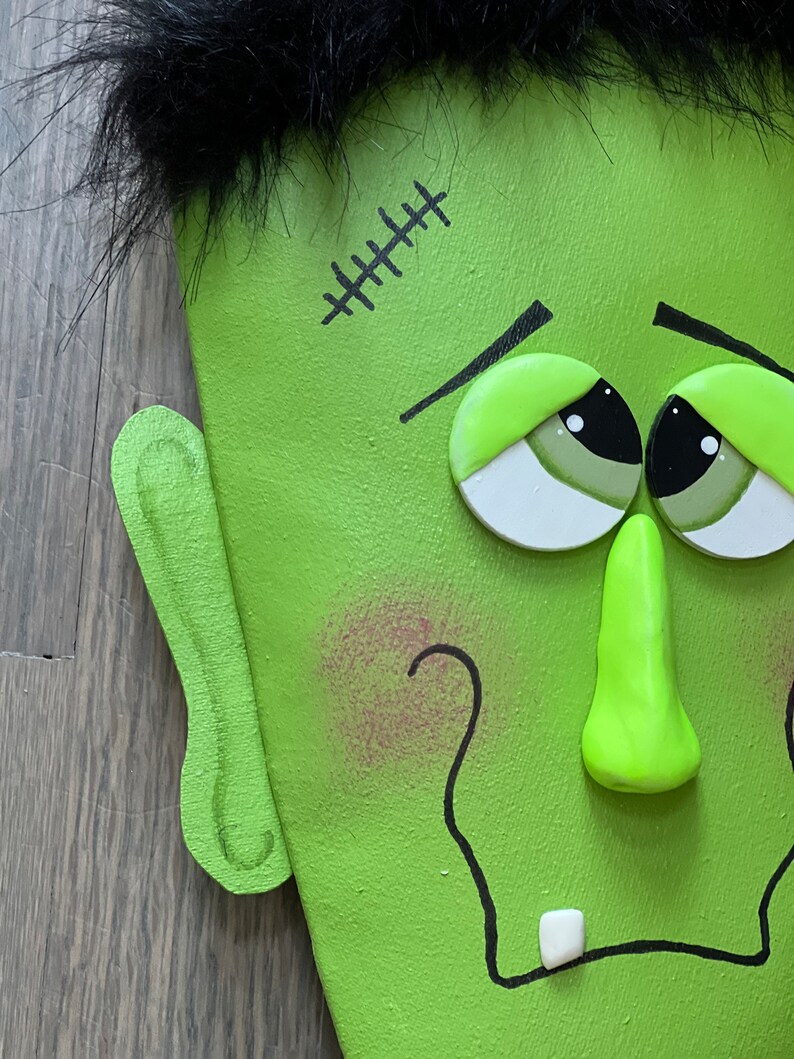 May include: Close-up of a green Frankenstein face with black hair, stitched scar, and a toothy grin. The face is made of wood and painted with details.