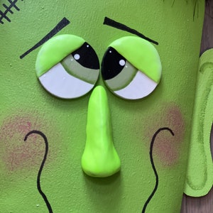 May include: A green monster face with black eyes, a green nose, and a stitched scar on the forehead. The monster has a white tooth sticking out of its mouth.