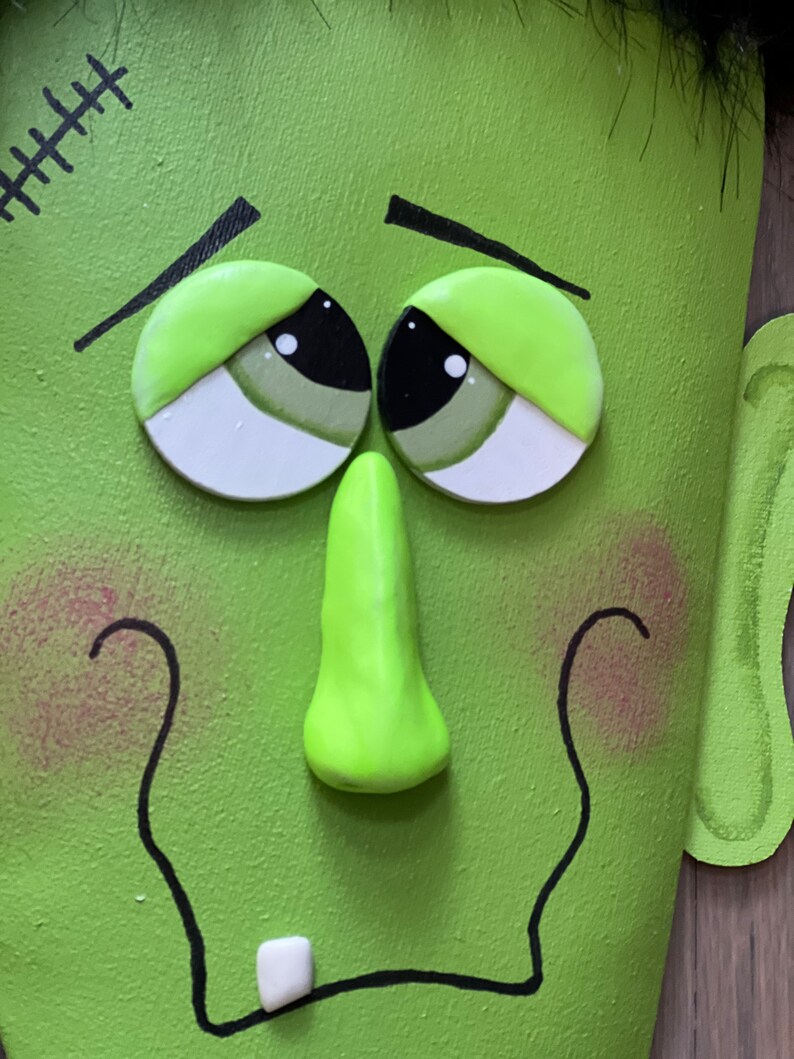 May include: A green monster face with black eyes, a green nose, and a stitched scar on the forehead. The monster has a white tooth sticking out of its mouth.