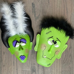 May include: A pair of green monster faces with black and white hair. The faces have stitched scars, googly eyes, and a toothy grin.