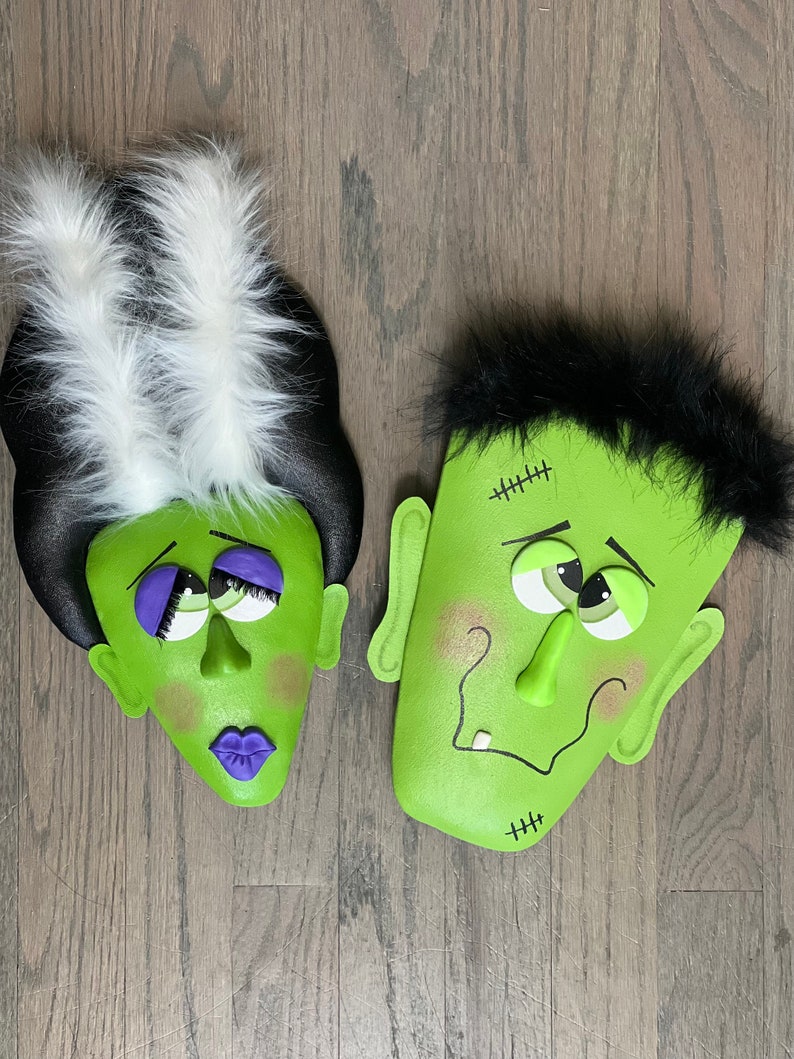 May include: A pair of green monster faces with black and white hair. The faces have stitched scars, googly eyes, and a toothy grin.