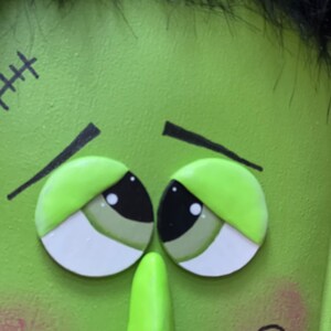 May include: Close-up of a green monster face with black hair, stitched scar, and large, sad eyes. The monster has a large, green nose and rosy cheeks.