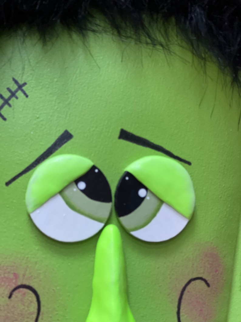 May include: Close-up of a green monster face with black hair, stitched scar, and large, sad eyes. The monster has a large, green nose and rosy cheeks.