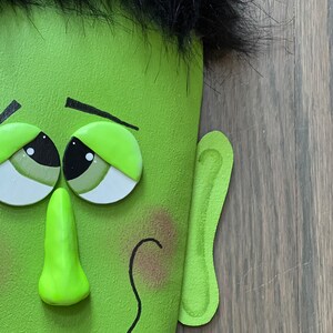 May include: A green Frankenstein-themed Halloween decoration with a black furry hairpiece, large green eyes, a green nose, and a crooked smile.