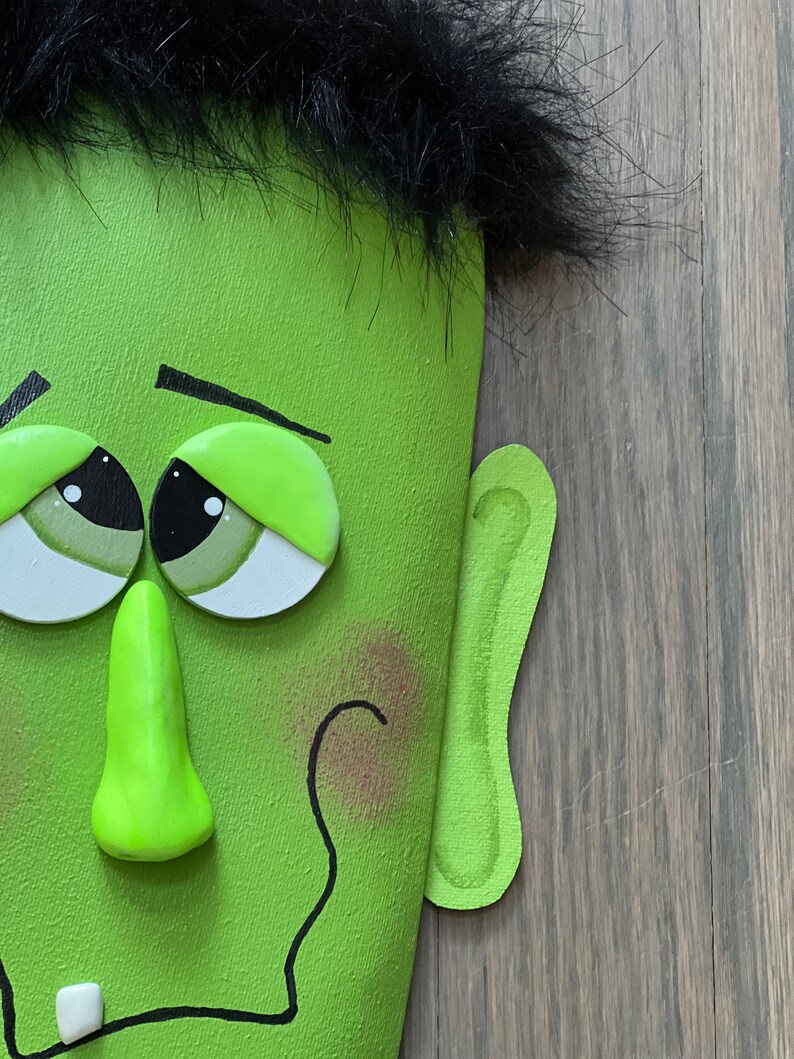 May include: A green Frankenstein-themed Halloween decoration with a black furry hairpiece, large green eyes, a green nose, and a crooked smile.