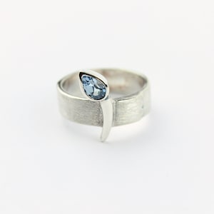 May include: A silver ring featuring a blue gemstone set in a pear shape. The ring band is textured and has a distinctive design.