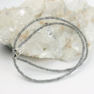 May include: A delicate silver chain necklace with small, gray labradorite beads. The necklace is displayed on a white crystal background.