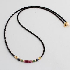 Könnte beinhalten: Eine schwarze Perlenkette mit einem bunten Edelstein-Akzent. Die Kette hat eine Reihe kleiner, bunter Edelsteine in den Farben Rosa, Blau, Grün und Gelb.