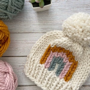 Op de afbeelding: Een witte gebreide muts met een regenboogontwerp en een pluche witte pompon bovenop. De regenboog is gemaakt van geel, roze en blauw garen.