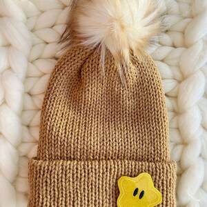 May include: A tan knit beanie with a folded brim and a large, fluffy, cream-colored pom-pom. A yellow star-shaped applique with black eyes is sewn on the brim. The hat is made of a thick, textured yarn.
