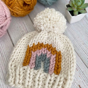 Op de afbeelding: Een witte gebreide beanie met een regenboogontwerp in tinten mosterdgeel, roze en grijs. De beanie heeft een pluche witte pompon bovenop.