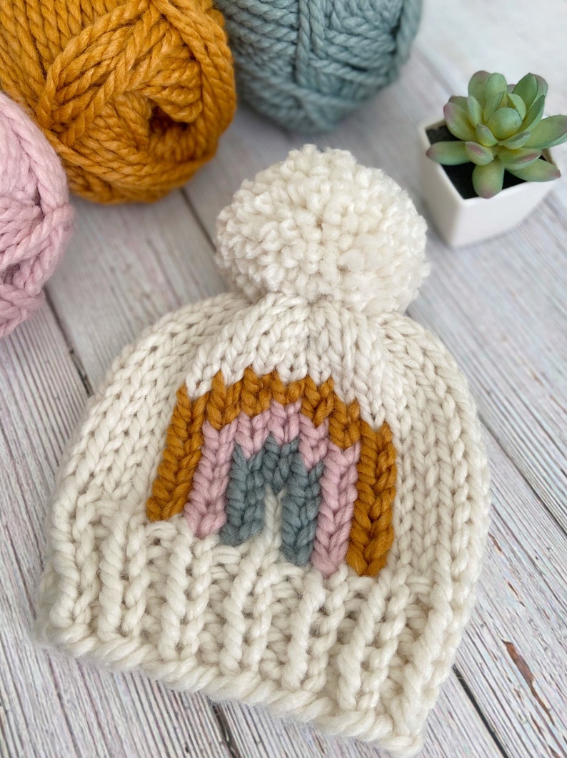 Op de afbeelding: Een witte gebreide beanie met een regenboogontwerp in tinten mosterdgeel, roze en grijs. De beanie heeft een pluche witte pompon bovenop.
