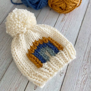 Op de afbeelding: Witte gebreide muts met een pompon bovenop en een regenboogontwerp in blauw, grijs en geel.